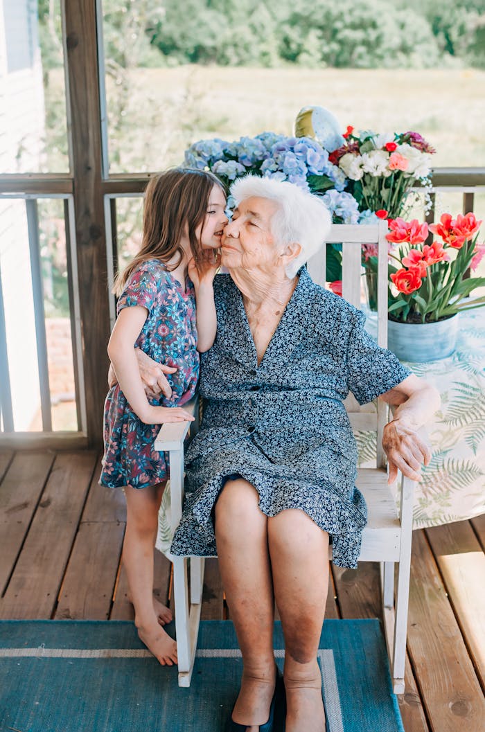A young girl kisses her grandmother on a porch, surrounded by colorful flowers.